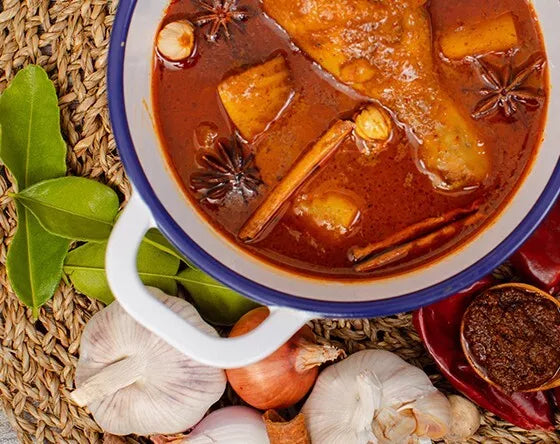 Delicious Thai Massaman curry served in a pot with chicken, potatoes, cinnamon sticks, star anise, and traditional spices, surrounded by fresh ingredients like garlic, onion, lemongrass, and curry paste.